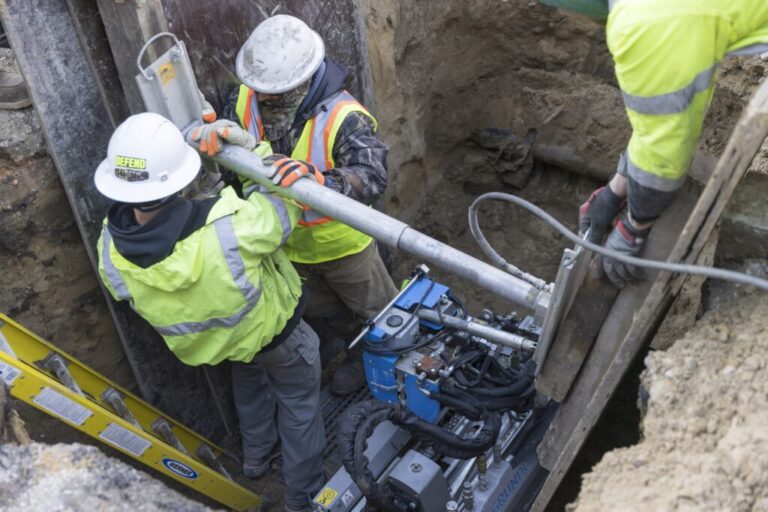 The photo show workers in hard hats and reflective vests in a trench with equipment.