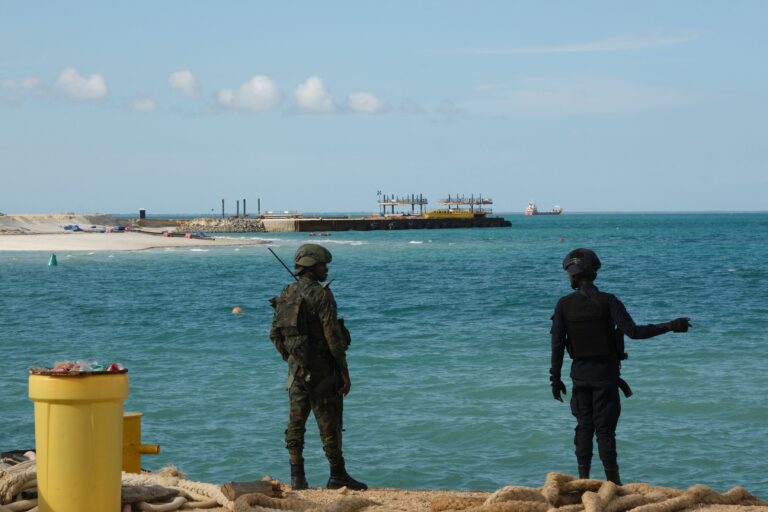 The police officer and solider are in the foreground; beyond them, water stretches out toward the project.